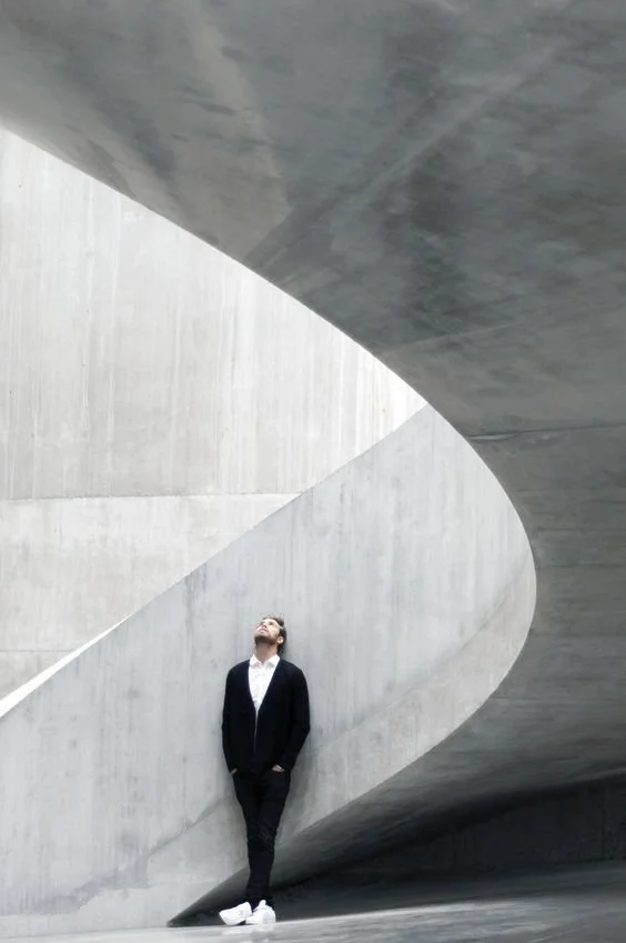 Persona de pie en una estructura de concreto moderna y minimalista, observando hacia arriba en un entorno arquitectónico abstracto.
